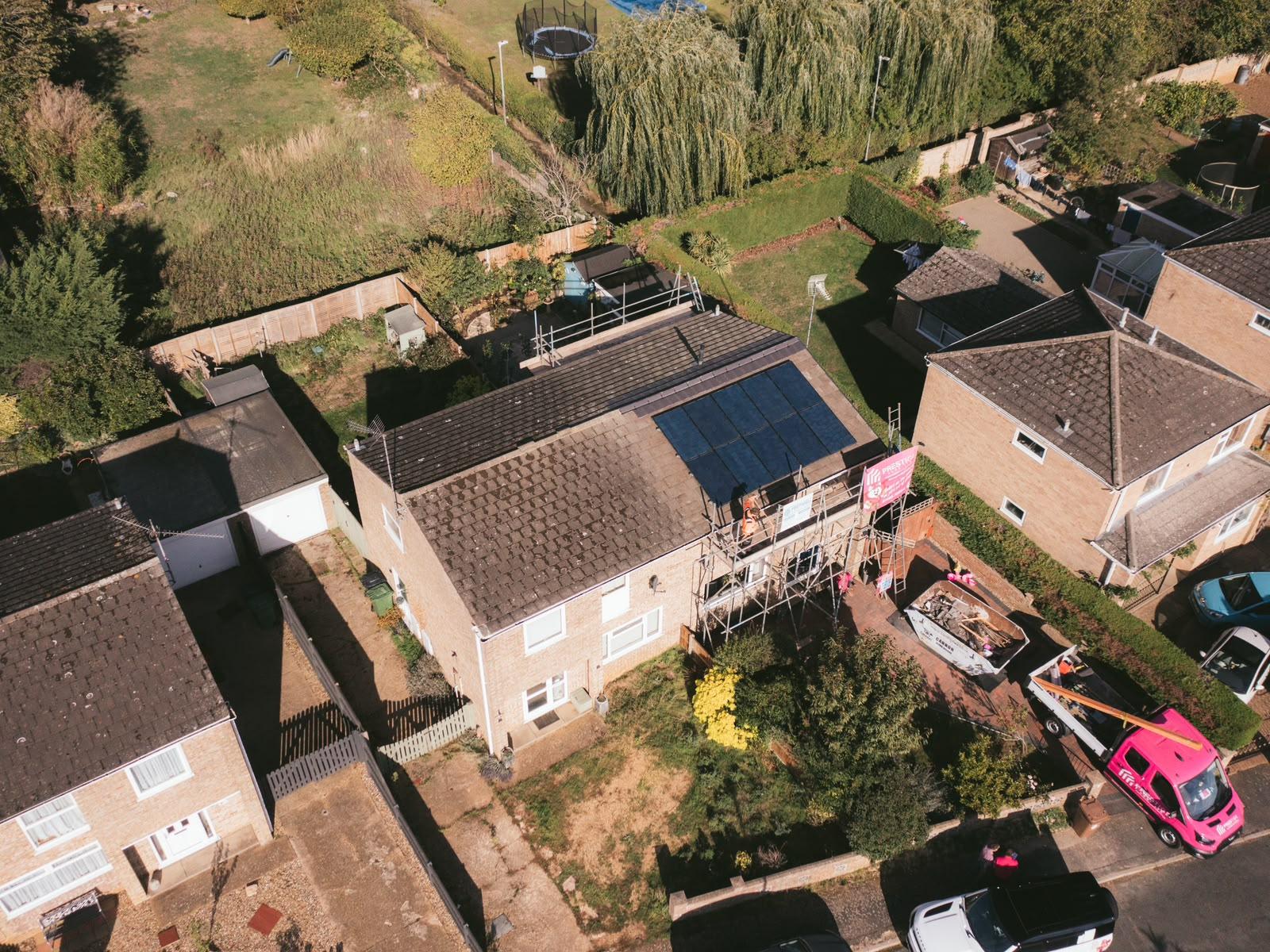 Wide aerial view of the street with the scaffolded house and a pink Prestige Roof van on the drive