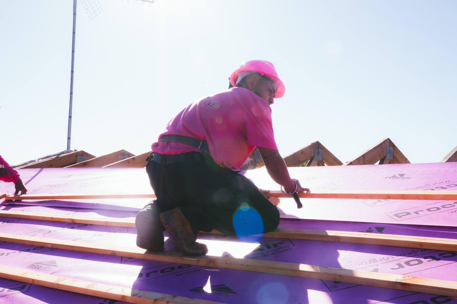 A roofer in a pink hard hat fixing battens on the roof in bright morning sunshine