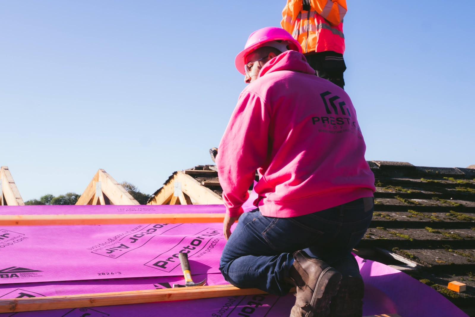 A Prestige Roof roofer in a pink hard hat and branded hoodie kneeling on the new roof membrane